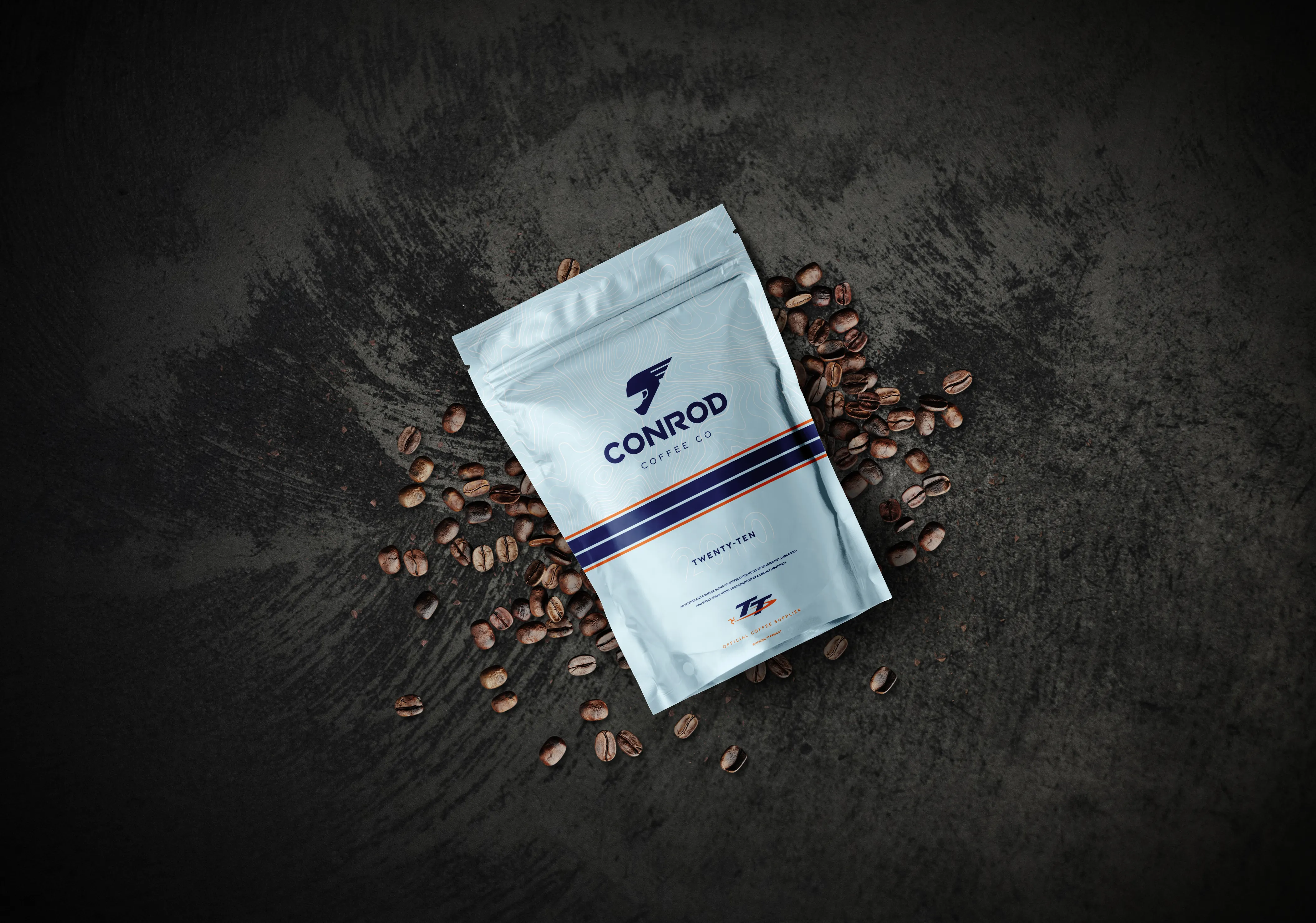 A white coffee bag labeled "Conrod Coffee Co" sits on a textured black surface, surrounded by scattered coffee beans, conveying a premium feel.